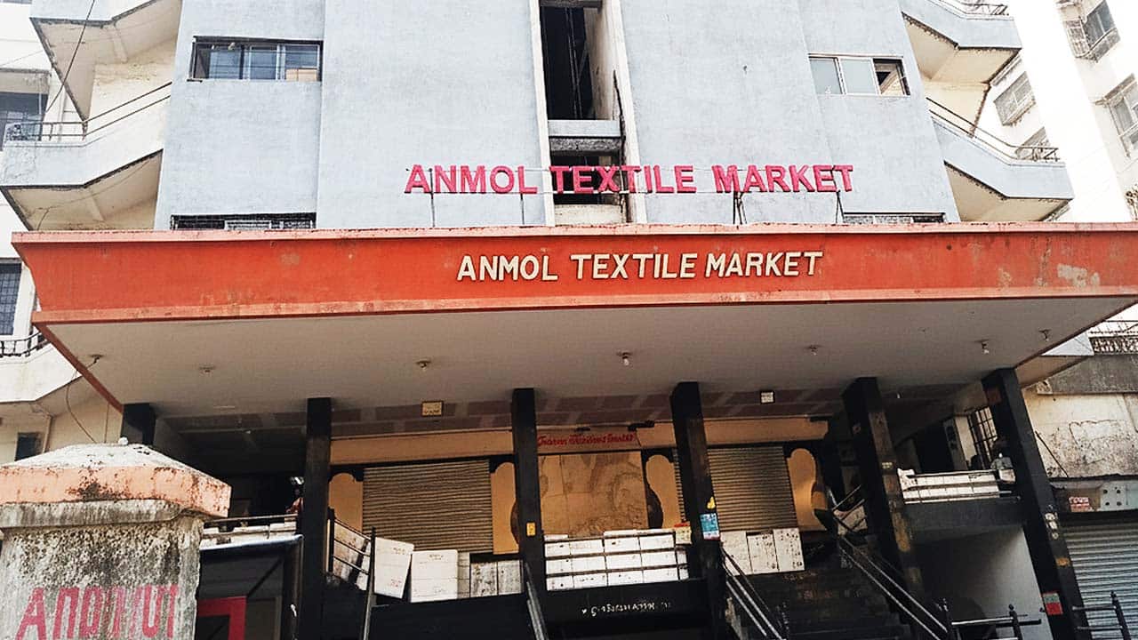The prominent red signage of "ANMOL TEXTILE MARKET" above the entrance of a multi-story market building in Surat.