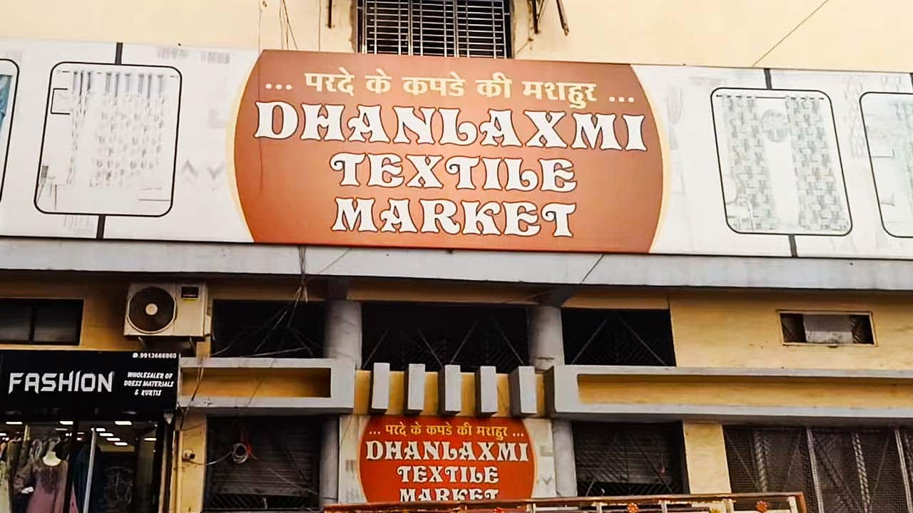 The prominent "DHANLAXMI TEXTILE MARKET" signage in large, bold lettering on a maroon background, with illustrations of curtains on either side.