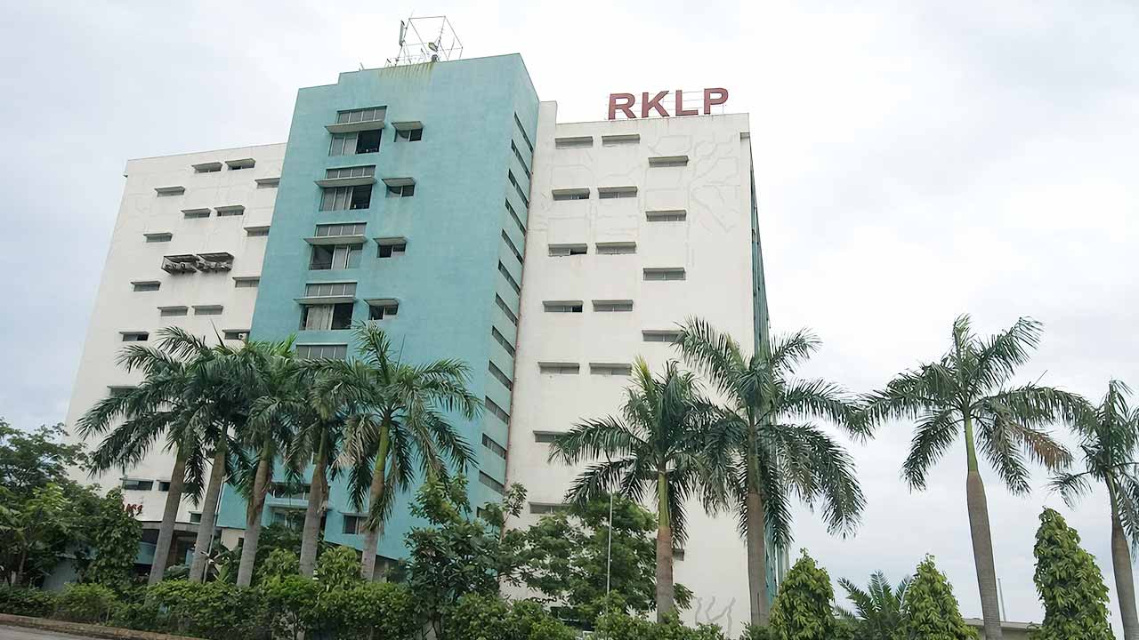 A multi-story building with a light blue and white facade, identified by the "RKLP" logo, surrounded by palm trees.
