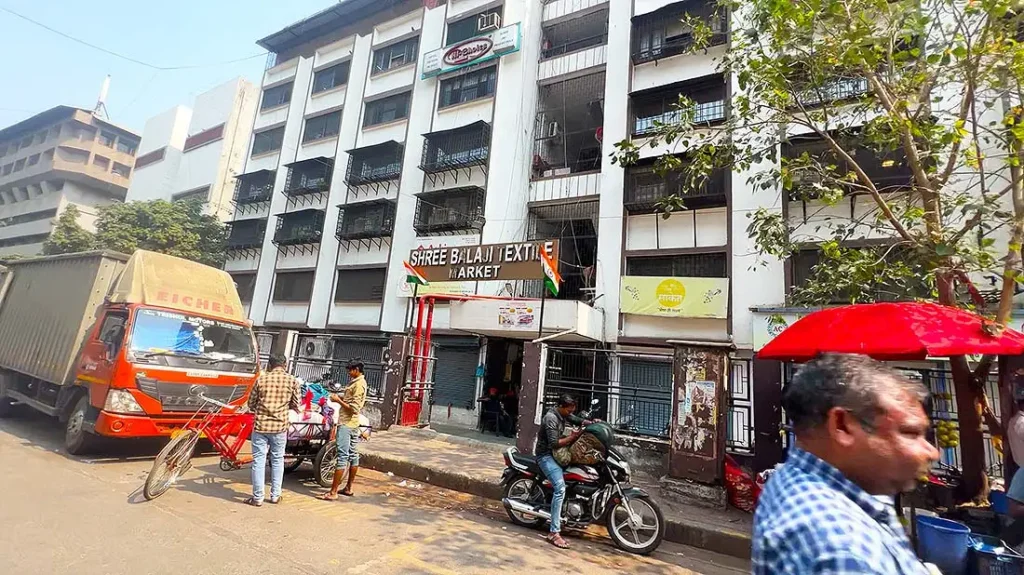 Wide view of Shree Balaji Textile Market building on Ring Road, Surat, with transport vehicles and wholesale textile buyers.